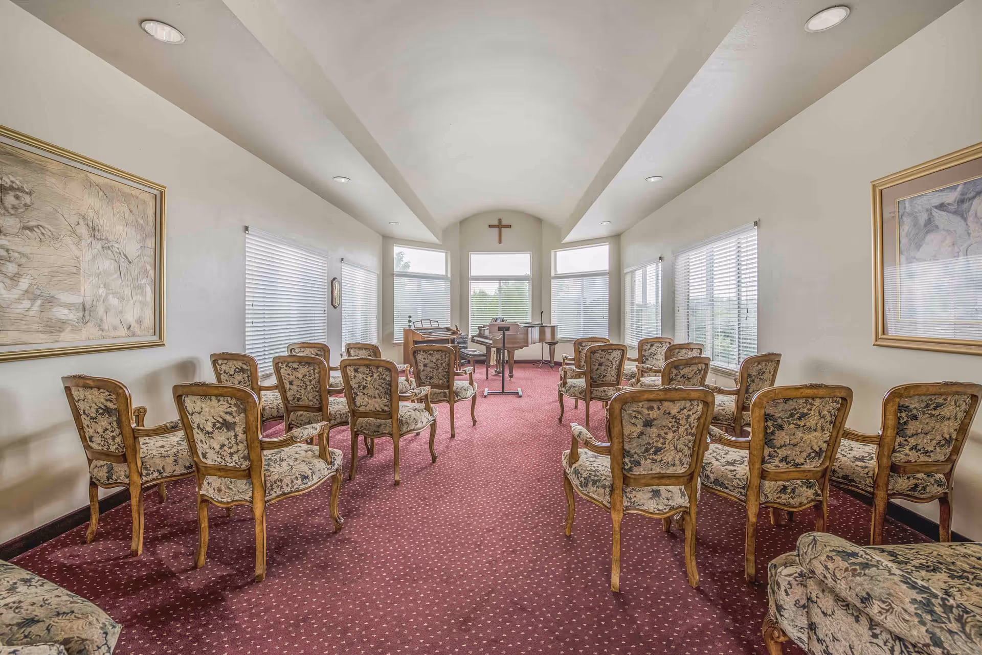 Room with rows of upholstered floral chairs on red carpet facing a piano and a cross on the wall, with windows along both sides.