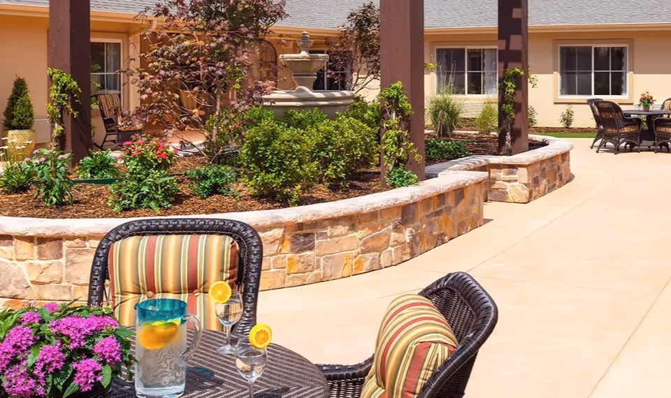 Sunlit courtyard with wicker patio seating and a table with drinks in front of a fountain and landscaped flowerbeds outside a memory care building.