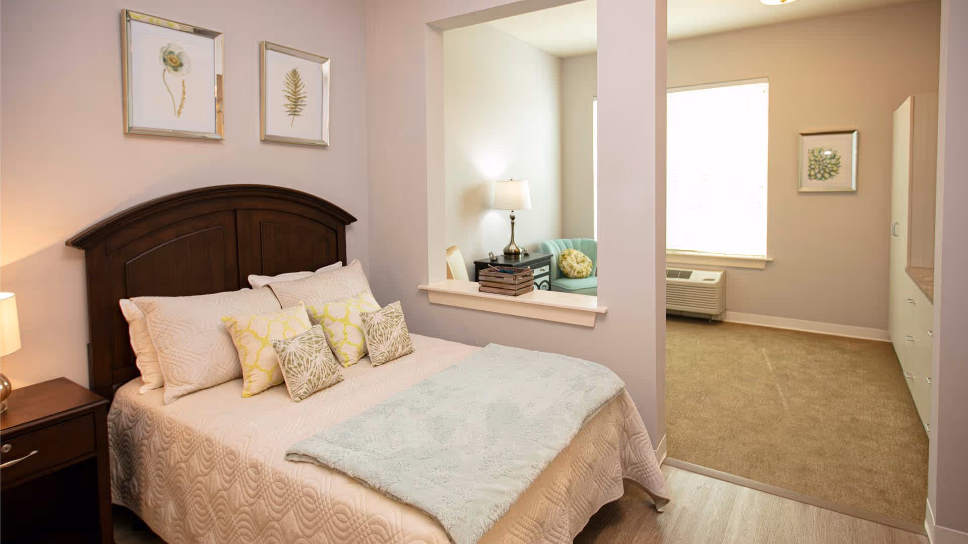 A cozy bedroom with a wooden headboard bed adorned with cream and green patterned pillows and a light blue throw blanket. The room has soft beige walls with two framed botanical prints above the bed. A nightstand with a lamp is beside the bed. There is a large open window-like cutout in the wall showing a small sitting area with a lamp, a side table, and a light green chair. The adjacent room has a window with blinds, a wall-mounted air conditioning unit, a framed botanical print, and a white cabinet with drawers.