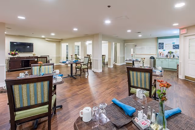 A spacious dining room in a senior living facility with wooden floors, several tables set with placemats, napkins, glasses, and small flower vases. There are upholstered chairs around the tables, a television mounted on the wall, and a counter area with coffee and other refreshments in the background. The room is well-lit with recessed ceiling lights.