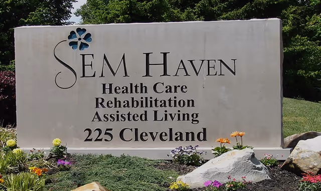 A large stone sign for SEM Haven displaying the services offered: Health Care, Rehabilitation, Assisted Living, and the address 225 Cleveland. The sign is surrounded by greenery, flowers, and rocks.