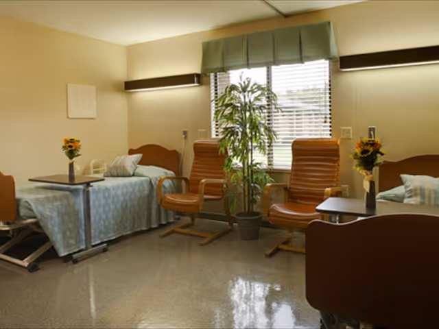 A senior living facility room with two hospital-style beds, each with a light blue blanket and a pillow. Between the beds are two brown leather chairs and a potted plant in front of a window with blinds and a green valance. Each bed has a small table with a vase of sunflowers.