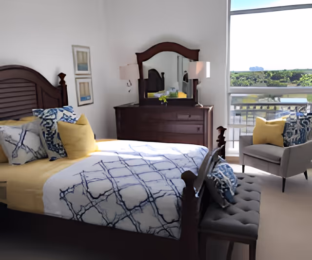 Sunlit bedroom with a wooden bed and patterned bedding, a dresser with mirror, and an armchair by a large window overlooking greenery.