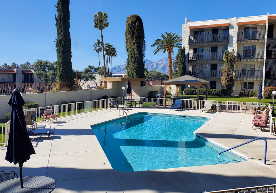 Outdoor swimming pool area at Fellowship Square Tucson with clear blue water, surrounded by a concrete deck with lounge chairs, tables with umbrellas, and a small gazebo. Tall palm trees and other greenery are visible around the pool, with a multi-story residential building in the background and mountains in the distance under a clear blue sky.