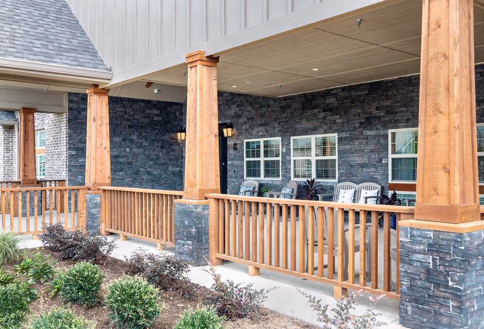 Covered front porch with wooden columns and railings, stone veneer wall, and outdoor seating at an assisted living facility.