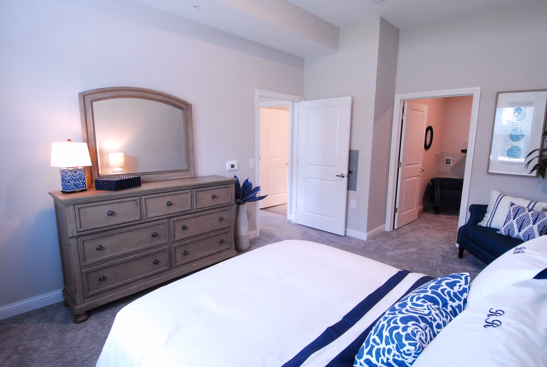Well-lit bedroom with a bed in the foreground, a dresser with mirror and lamp on the left, and open doors leading to adjacent rooms.