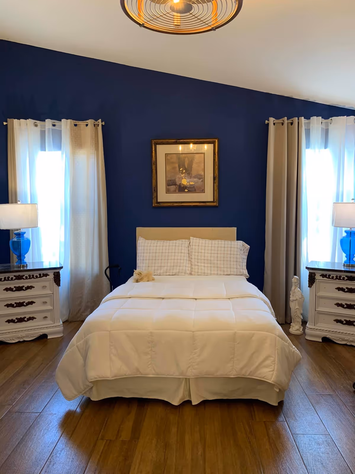 A neatly made bed with white bedding and checkered pillows is centered against a dark blue wall. On either side of the bed are white nightstands with ornate dark handles, each topped with a blue glass lamp with white shades. Two windows with white and beige curtains flank the bed. Above the bed hangs a framed picture. The floor is wooden, and a small statue is placed near the right nightstand.