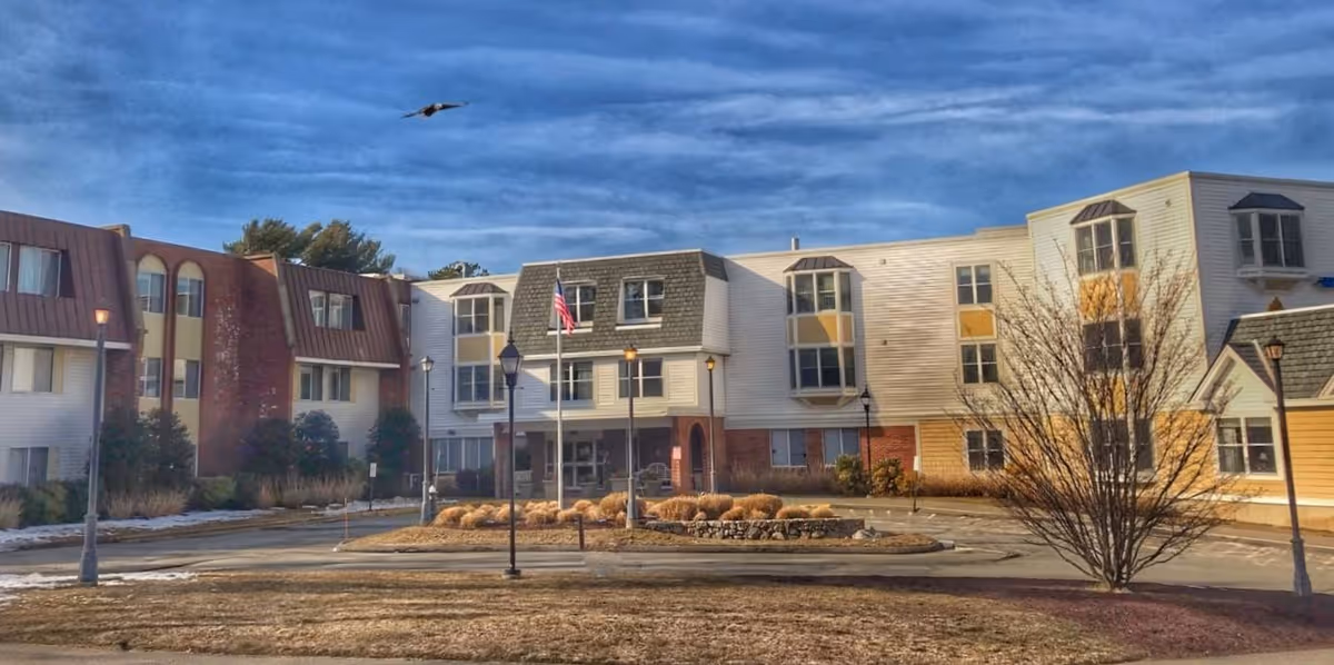 Exterior view of a multi-story senior living facility building with a circular driveway, several lamp posts, an American flag, and a leafless tree in front under a partly cloudy blue sky.