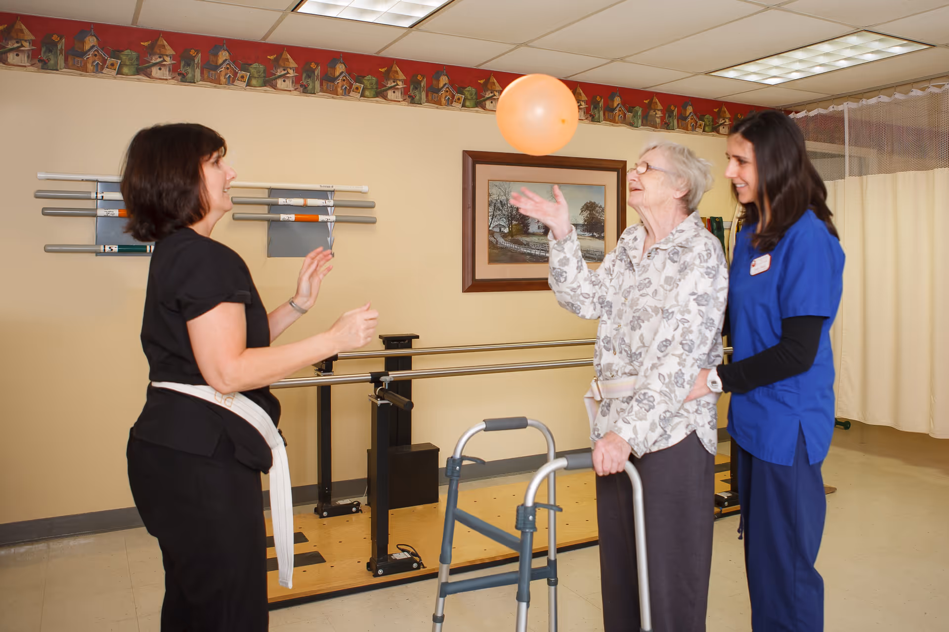 An elderly woman using a walker is playing with an orange balloon with the assistance of two female caregivers in a rehabilitation or therapy room. The room has handrails along the wall and a framed picture hanging above them. One caregiver is wearing black scrubs and the other is in blue scrubs, both smiling and engaged with the elderly woman.