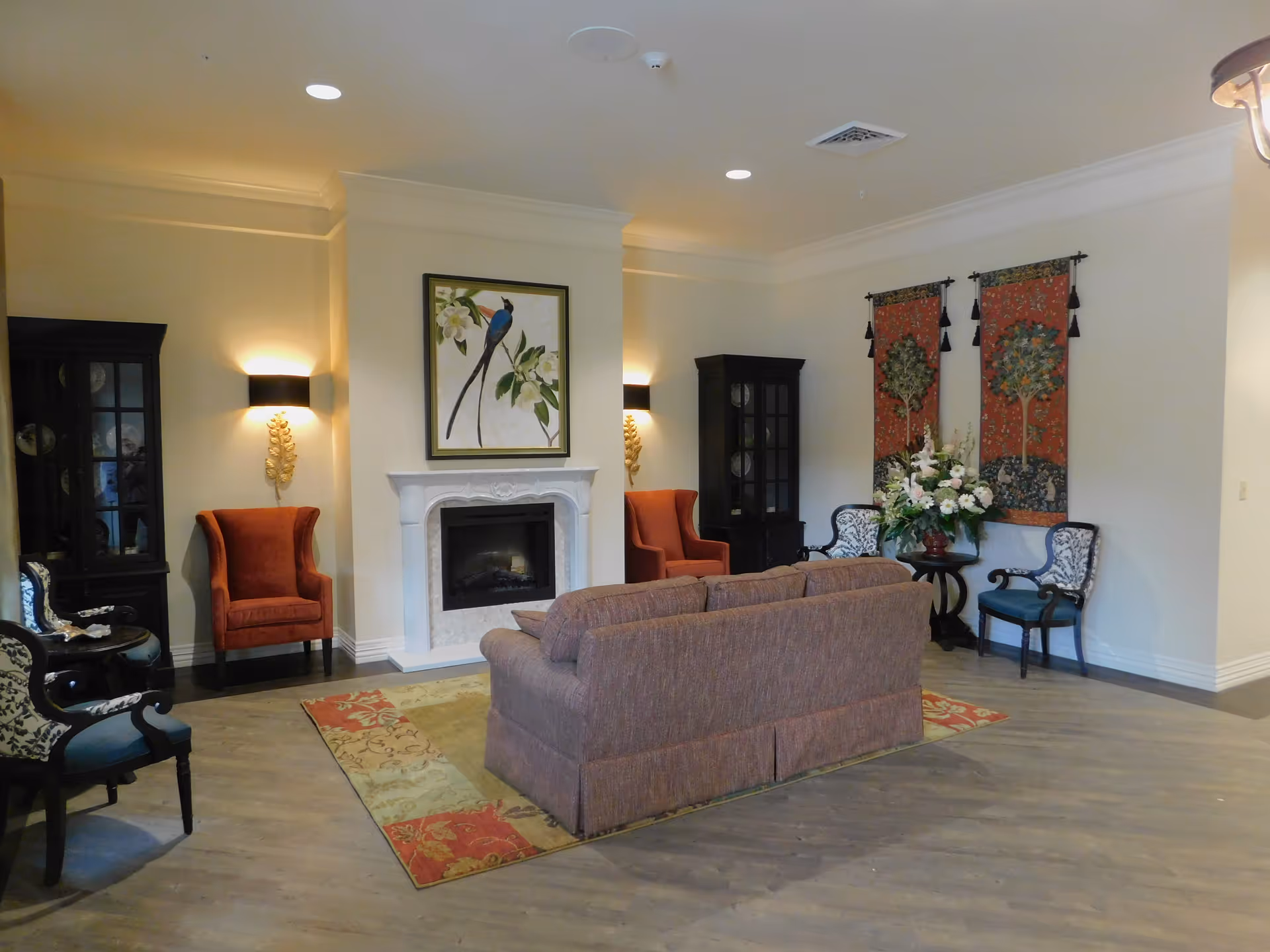 A cozy living room area with a brown sofa placed on a patterned rug facing a white fireplace. Above the fireplace is a framed painting of a bird on a branch. On either side of the fireplace are dark wood cabinets and orange armchairs. To the right, there are two decorative wall hangings and a small round table with a large floral arrangement. The room has wooden flooring and soft lighting from wall sconces.