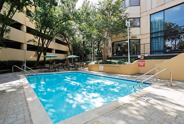 Outdoor swimming pool area surrounded by trees and patio furniture with umbrellas at Five Star Premier Residences Of Dallas. The pool is rectangular with metal handrails and a sign indicating a depth of 3 feet. The building exterior and a parking garage are visible in the background.