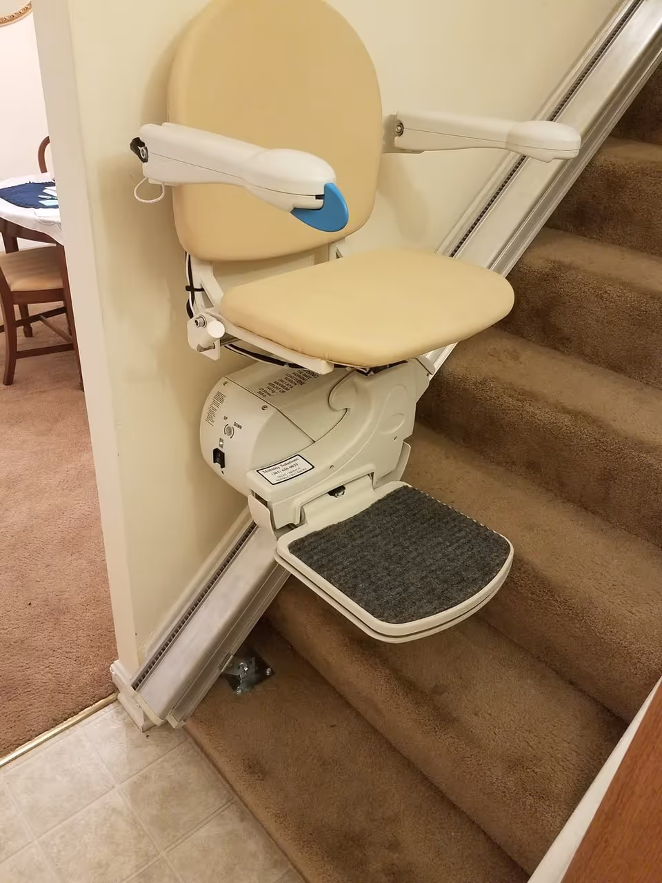 A beige stairlift installed on a carpeted staircase with a padded seat, armrests, and a footrest. The stairlift is mounted on a metal rail attached to the side of the stairs. In the background, part of a dining area with a wooden chair and table is visible.