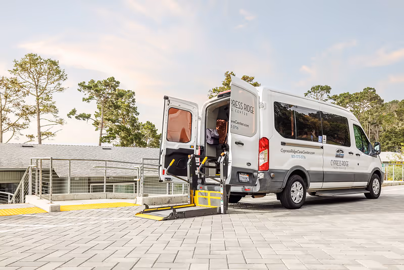A white van with the rear doors open and a wheelchair lift extended, parked on a paved area outside a building with trees in the background. The van has signage for Cypress Ridge Care Center.