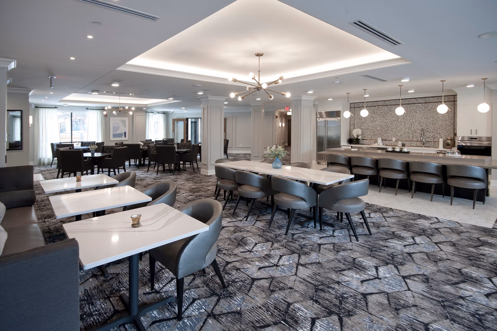 Spacious and modern dining area with multiple tables and chairs, a long marble table with chairs, and a kitchen area with a large island and bar stools. The room features patterned carpet, pendant lighting, and large windows with curtains allowing natural light.