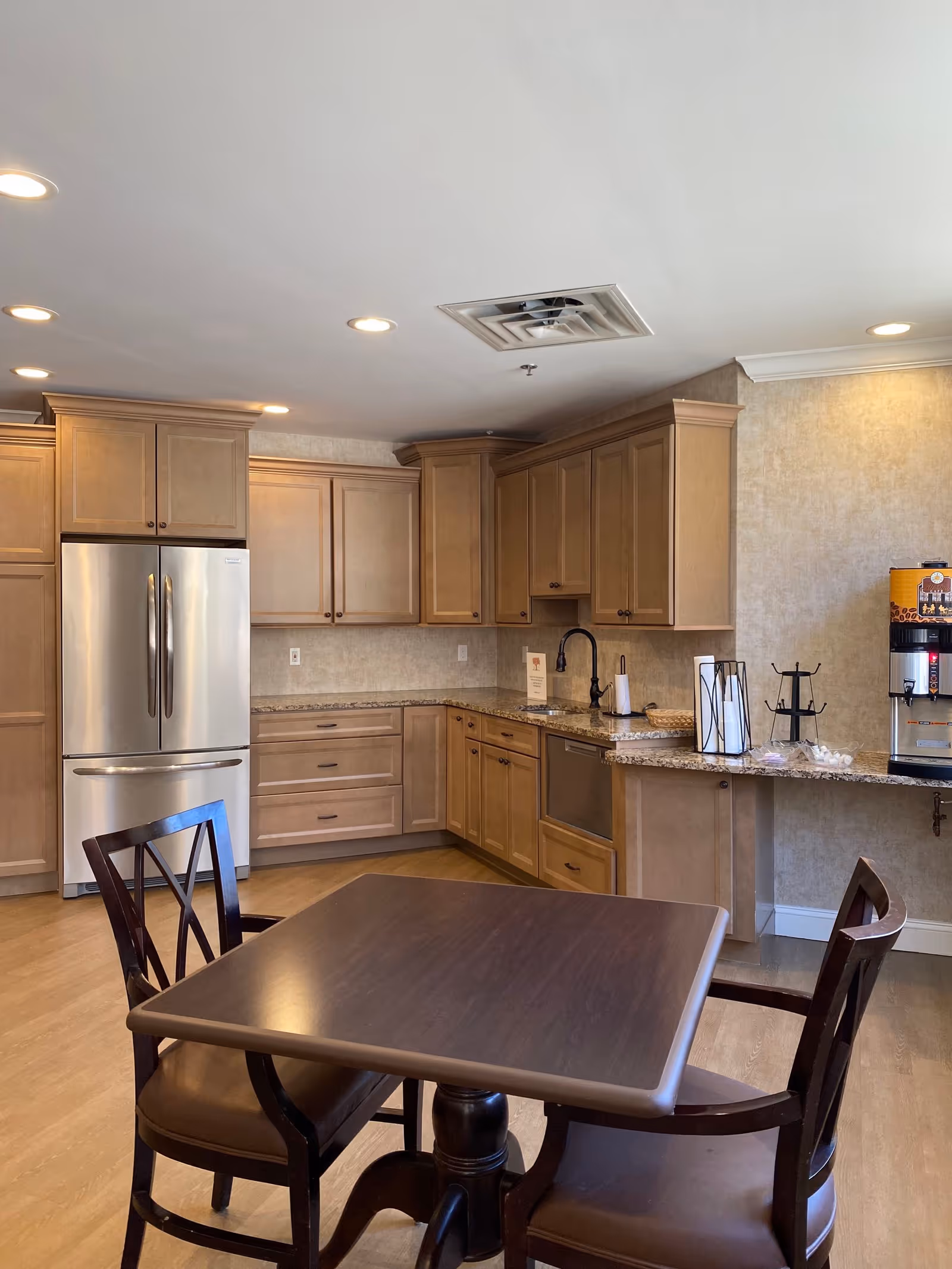 A communal kitchen and dining area with wooden cabinets, a stainless-steel refrigerator, granite countertops and a small wooden table with chairs.