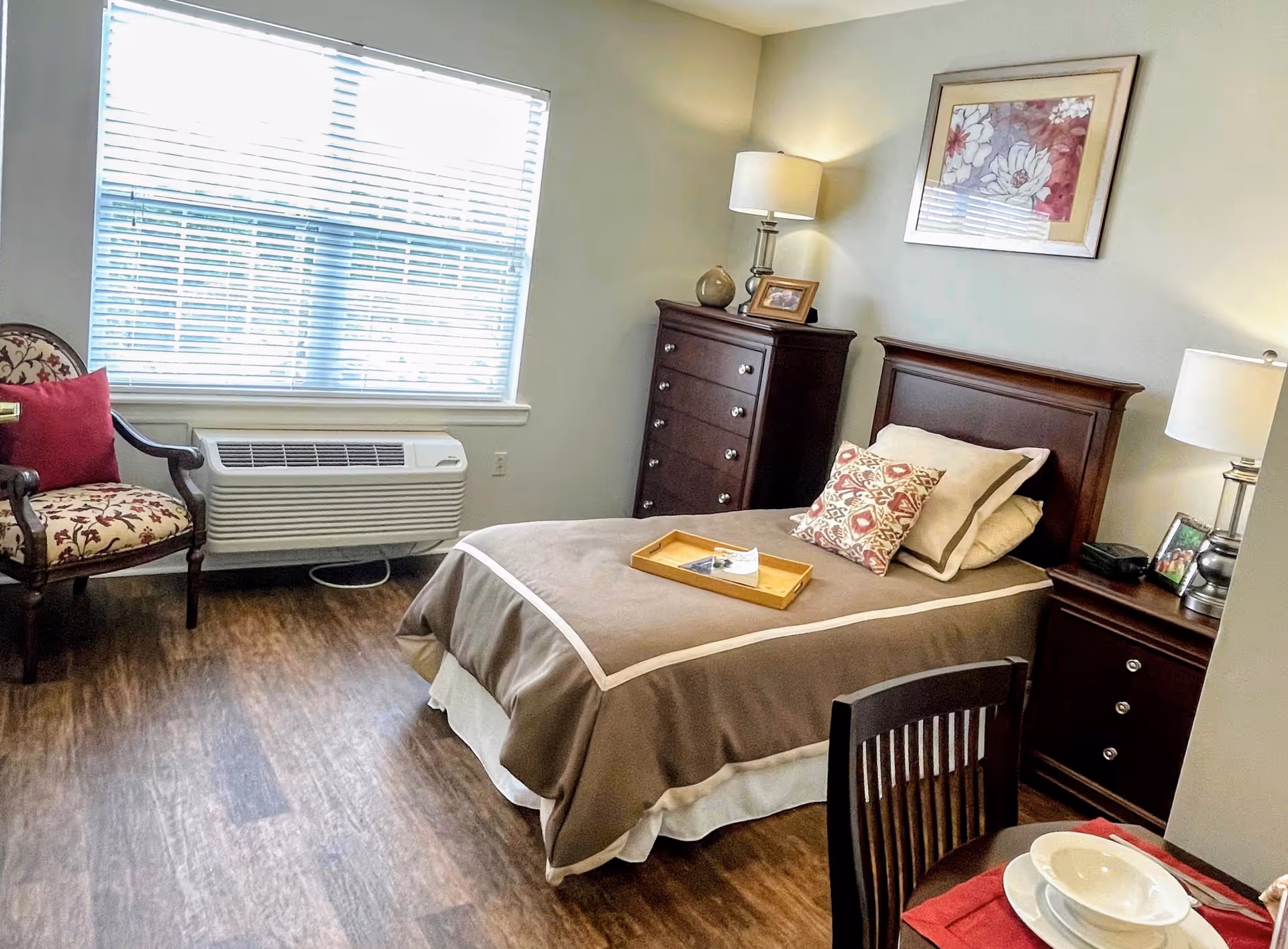 A cozy bedroom in a senior living facility featuring a single bed with brown and beige bedding, a wooden headboard, and decorative pillows. Next to the bed is a dark wooden nightstand with a lamp and framed photo. A matching tall dresser with a lamp and decorative items is against the wall. A window with blinds lets in natural light, and a patterned armchair with a red pillow sits nearby. The room has wood flooring and light-colored walls with a floral framed picture hanging above the bed.
