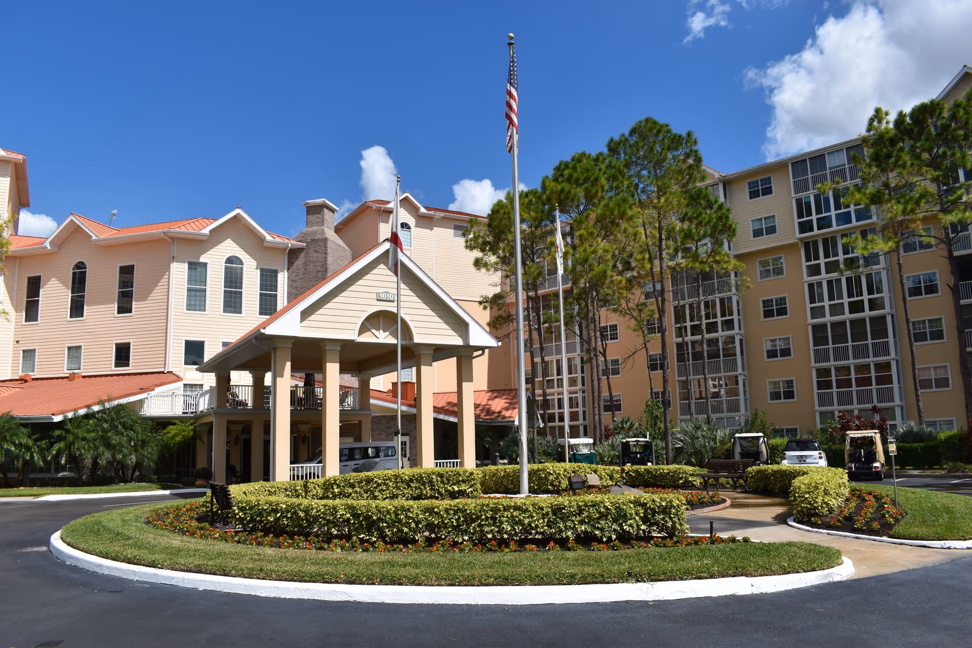 Circular landscaped driveway and flagpoles leading to the covered entrance of a multi-story senior living building.