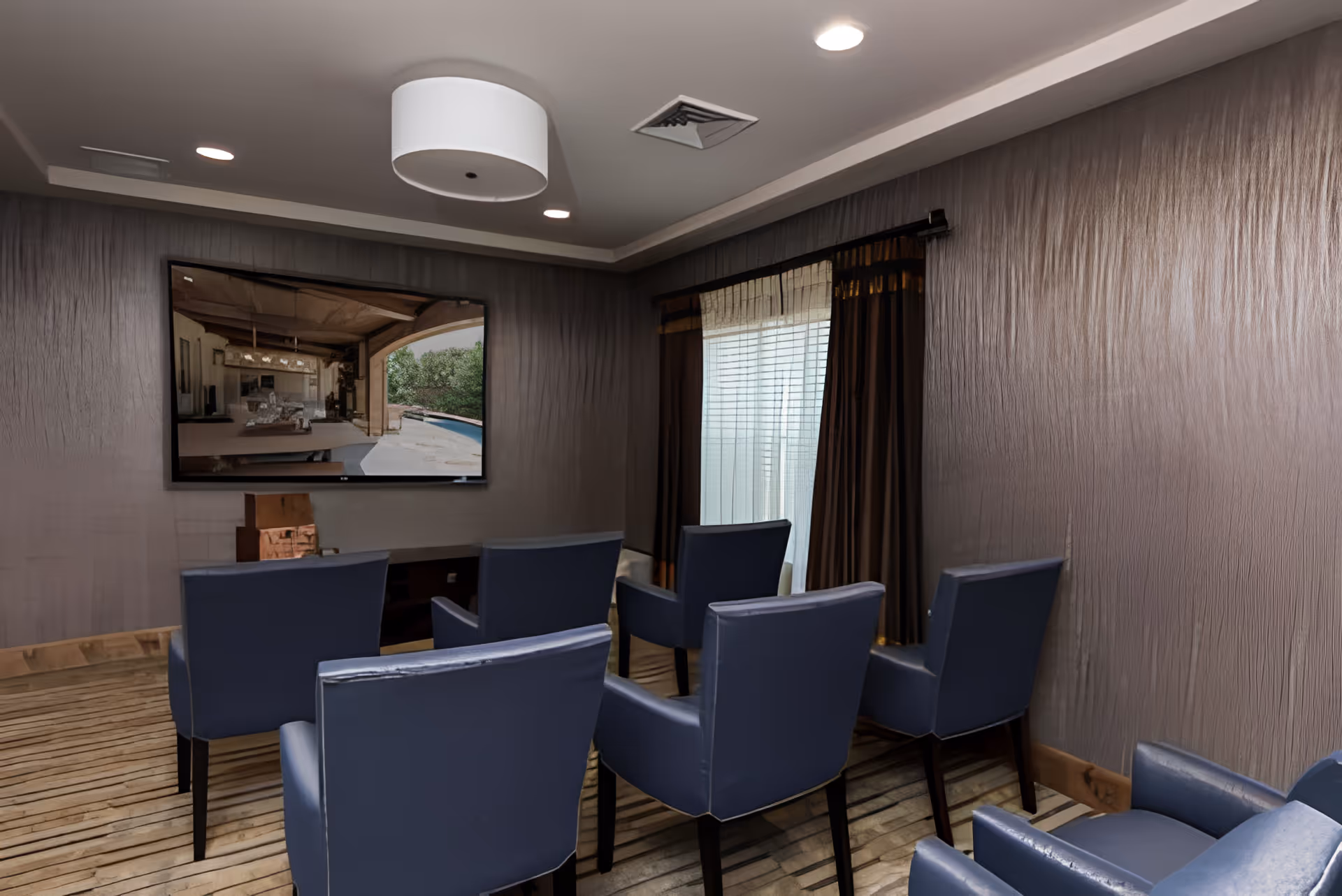 A small media room with six blue armchairs arranged in two rows facing a wall-mounted flat-screen TV. The room has textured gray walls, a wooden floor, a window with sheer and dark curtains, and a modern ceiling light fixture.