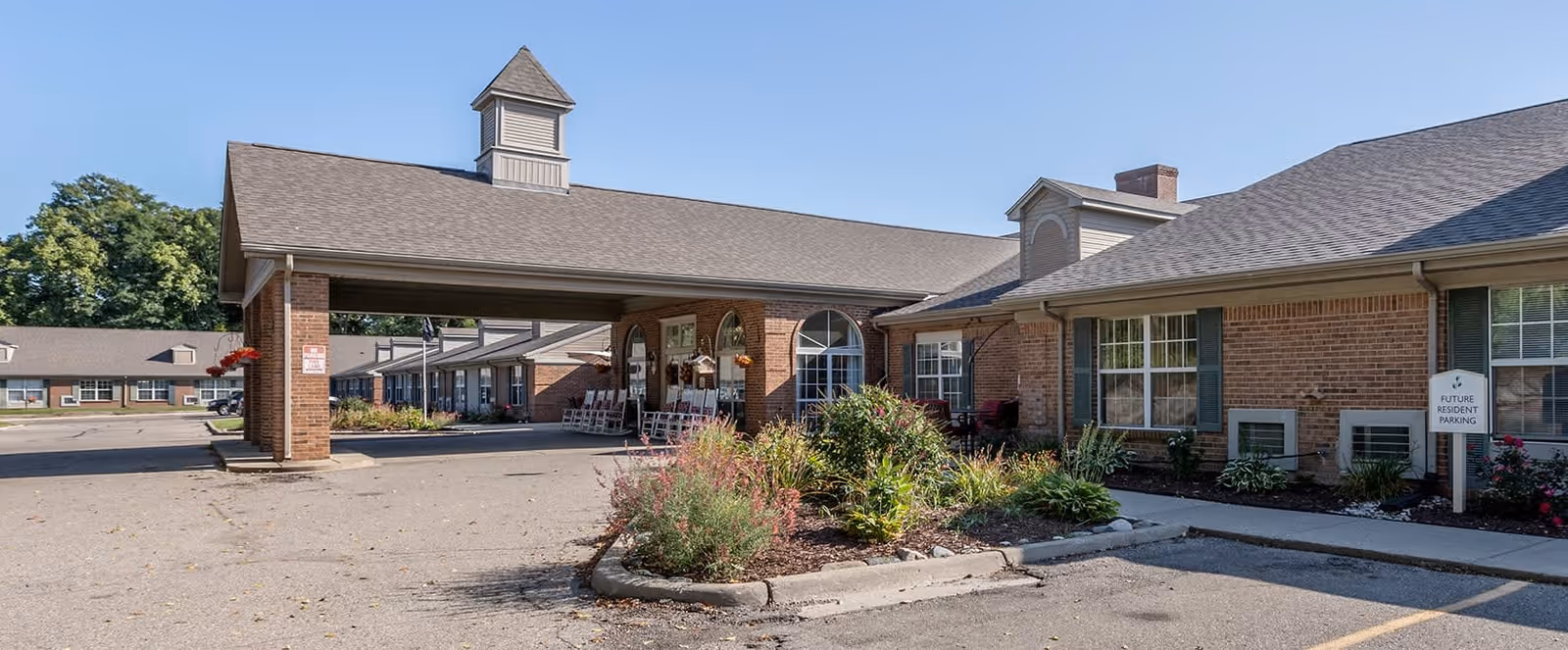 Exterior view of a single-story brick senior living facility with a covered entrance and a small garden area in front. The building has multiple windows with green shutters and a sign indicating future resident parking. The sky is clear and blue.