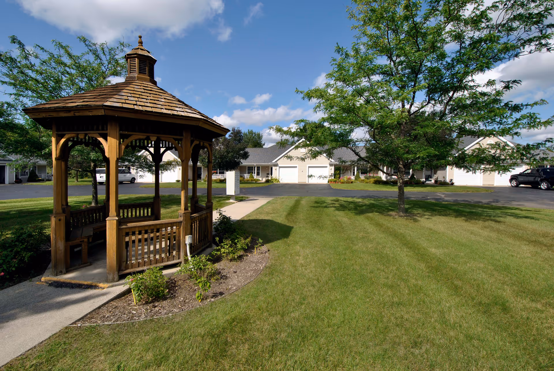 A wooden gazebo with a shingled roof situated on a concrete path surrounded by grass and small plants. In the background, there are trees, a paved driveway, and single-story residential buildings under a partly cloudy blue sky.
