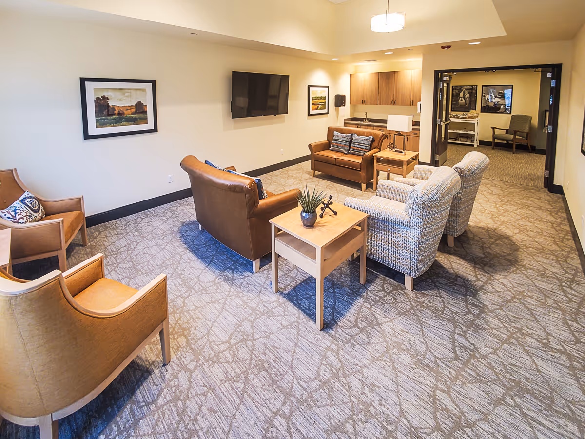 A cozy common lounge with armchairs, sofas, a wall-mounted TV, and side tables in an assisted living facility.