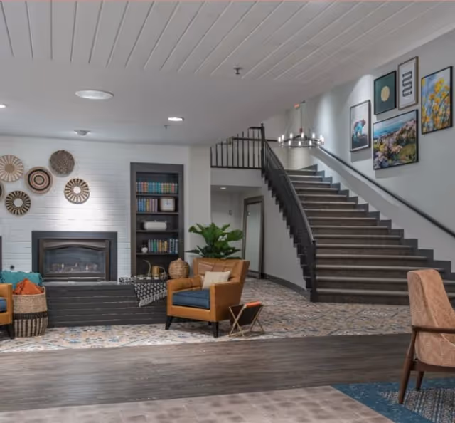 A cozy senior living facility common area featuring a fireplace with decorative wall hangings above it, a built-in bookshelf filled with books and decor, a comfortable armchair with a small side table, a large green plant, and a staircase with framed artwork on the wall leading to an upper floor.
