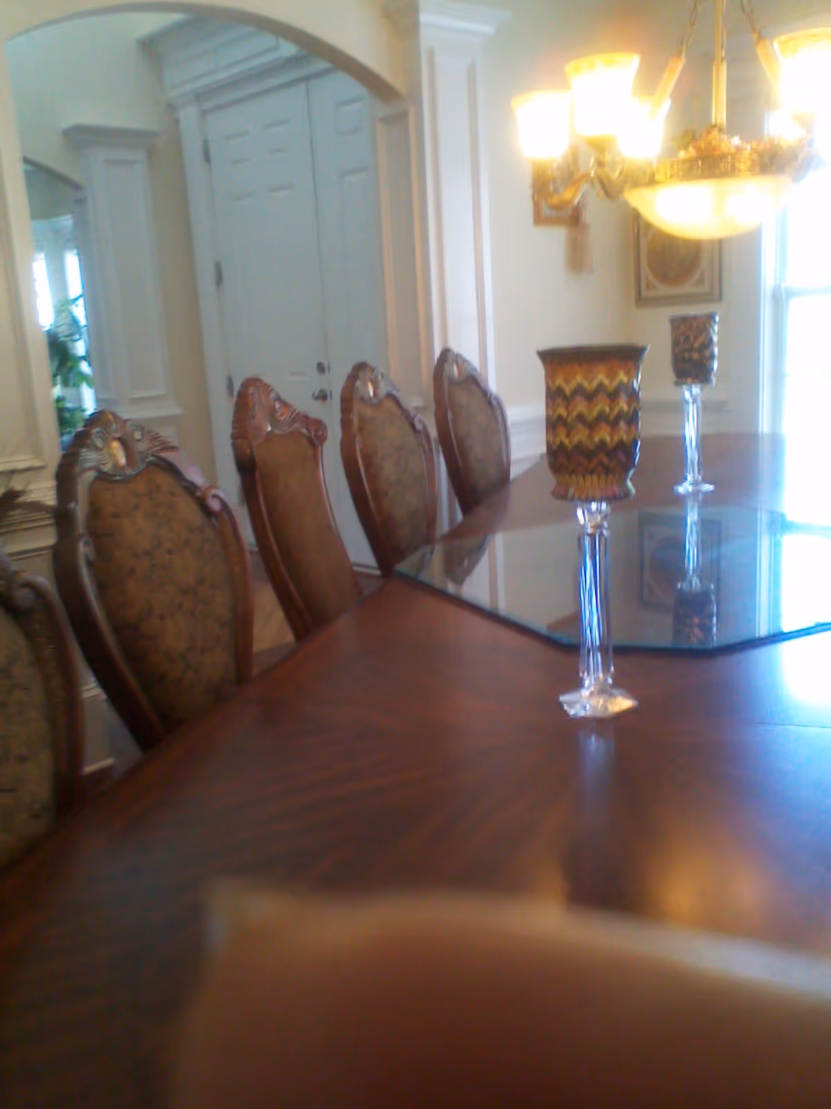 A formal dining room with a polished wooden table and six ornate chairs with patterned upholstery. On the table are two decorative candle holders with colorful designs. The room has white walls with molding, a chandelier with multiple lights, and a door in the background.
