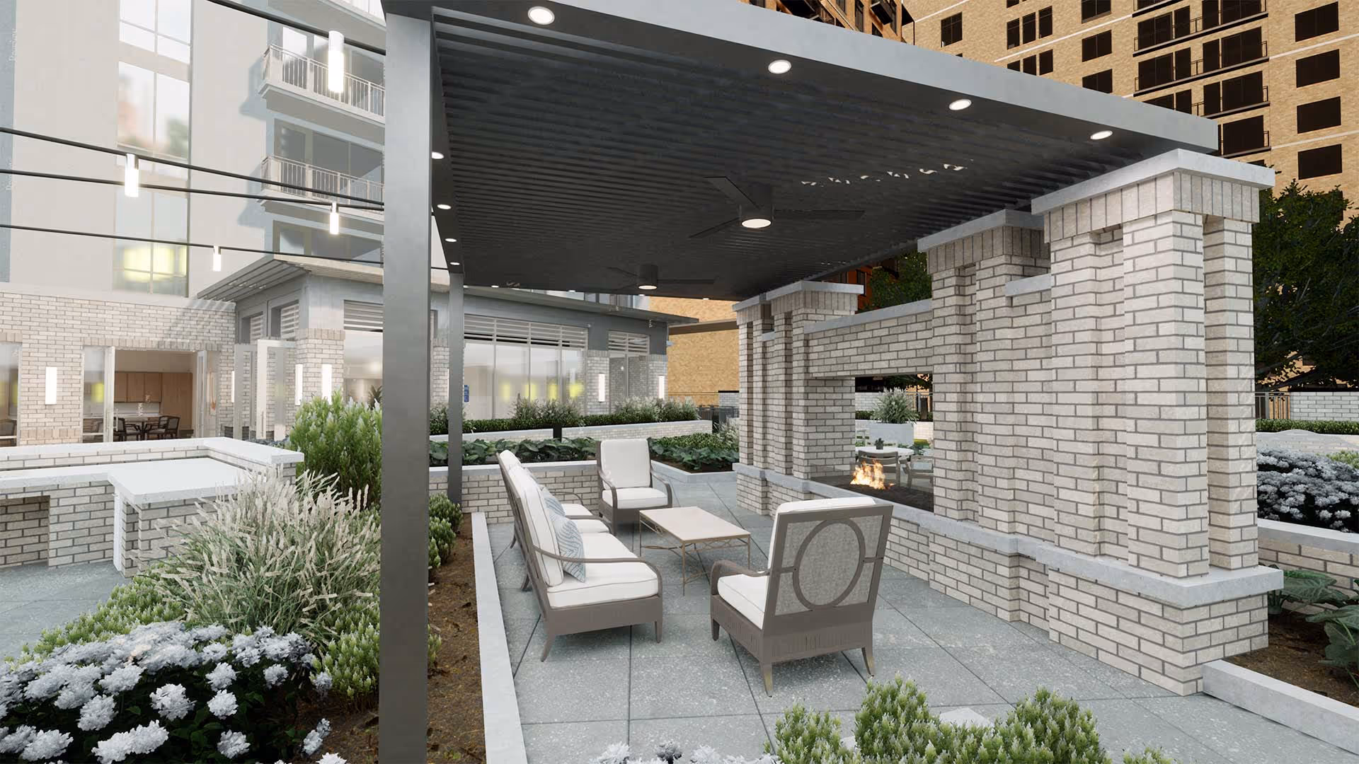 Outdoor seating area with cushioned chairs and a small table under a covered patio next to a modern brick fireplace, surrounded by landscaped greenery and buildings in the background.