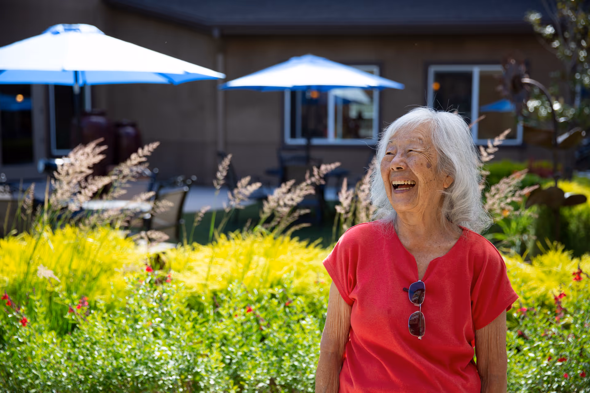 An elderly woman with white hair wearing a red shirt and sunglasses hanging from her collar is smiling and looking to the side while sitting outdoors in a garden area with green plants and flowers. In the background, there are patio tables with blue umbrellas and a building with windows.