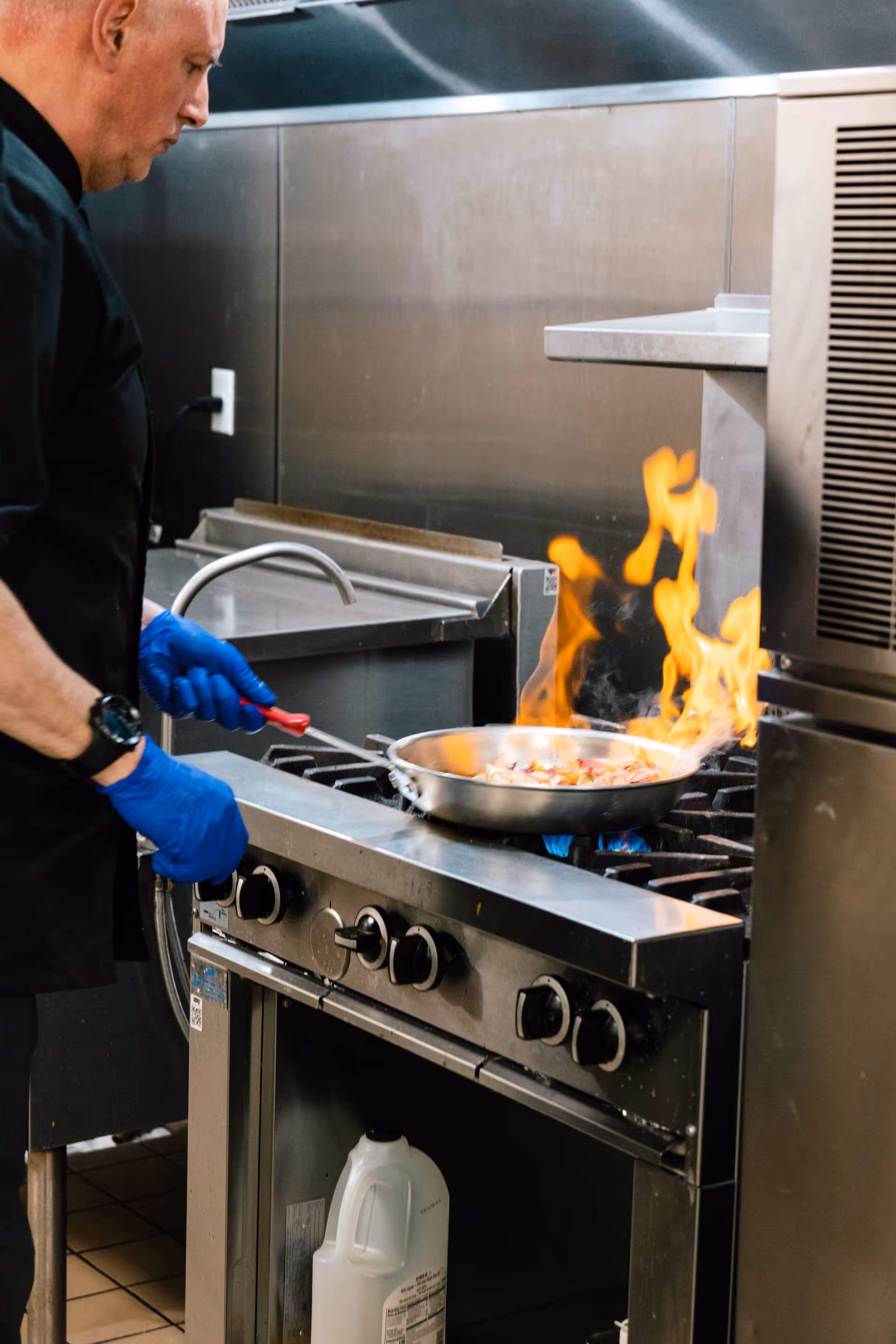 A chef wearing blue gloves is cooking food in a flaming pan on a commercial stove in a professional kitchen.