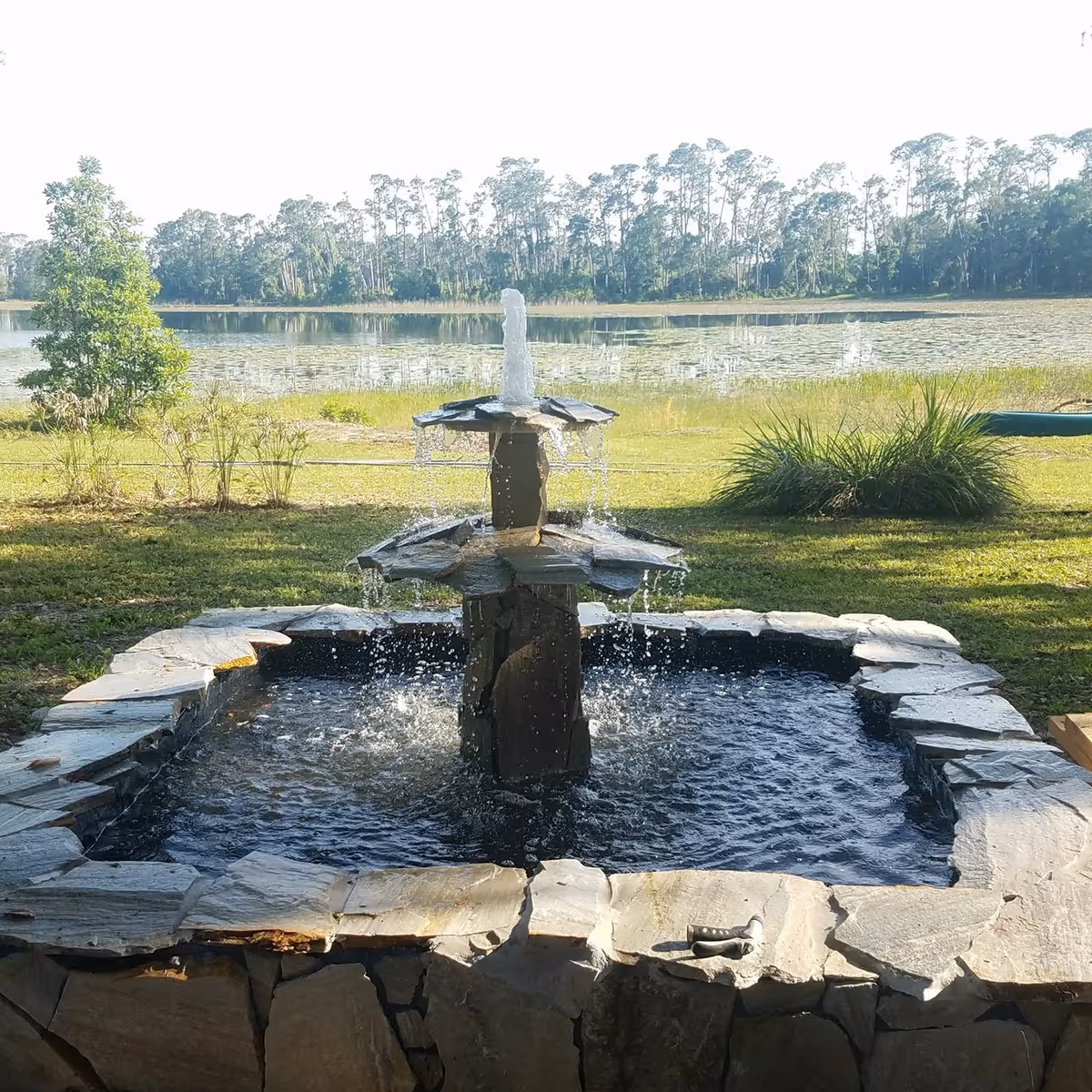 A stone water fountain with multiple tiers is flowing water, set in a square stone basin. The fountain is outdoors on a grassy area with a lake and trees in the background under a clear sky.