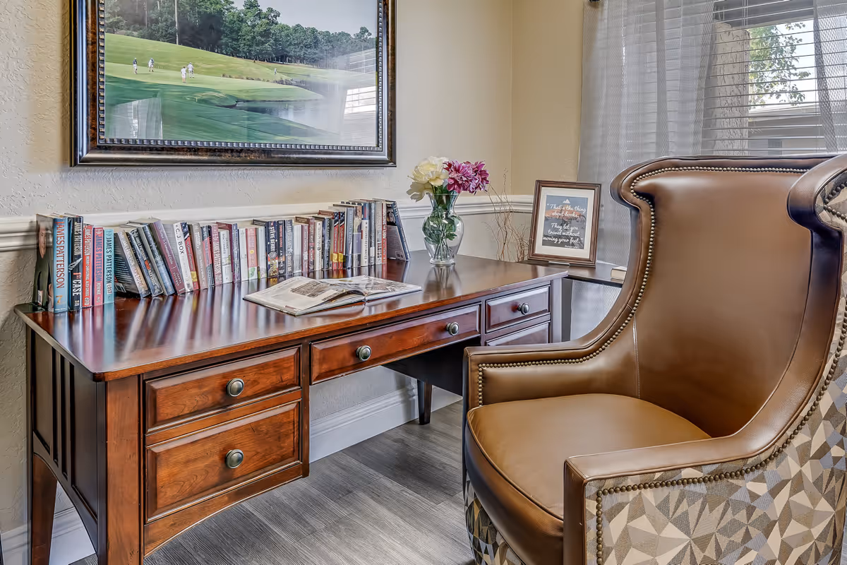 A cozy reading nook featuring a wooden desk with drawers, a collection of books lined up on the desk, an open book, a vase with flowers, and a framed inspirational quote. Next to the desk is a comfortable leather and fabric armchair. A large framed picture of a golf course hangs on the wall above the desk, and a window with sheer curtains allows natural light into the room.