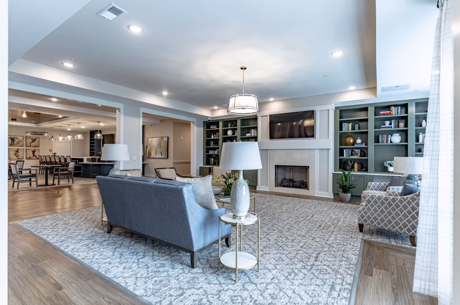 Spacious communal living room with sofas and chairs arranged around a fireplace and TV, built-in shelving, and a dining area visible in the background.