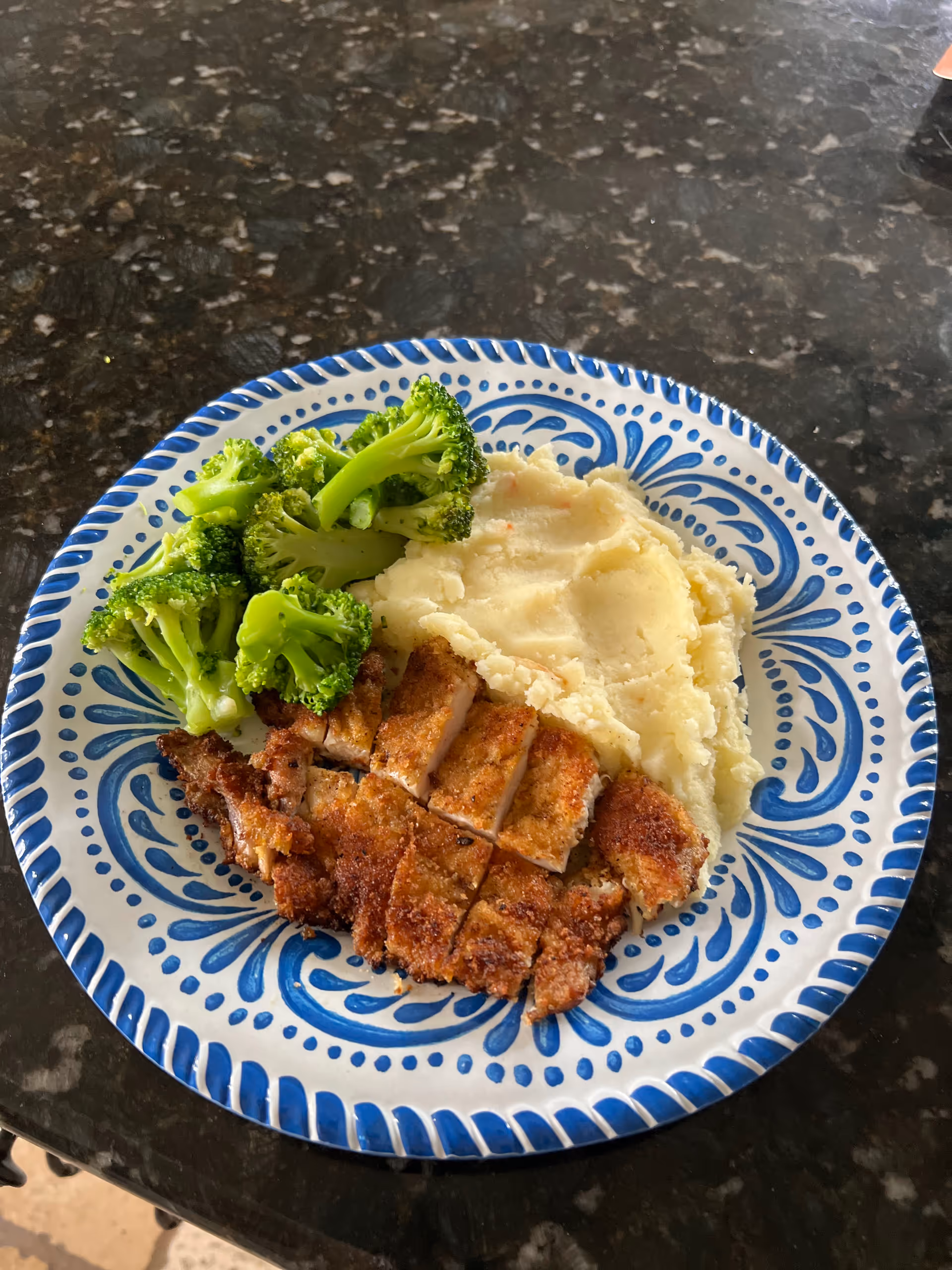 A plate with a serving of steamed broccoli, mashed potatoes, and sliced breaded chicken on a blue and white patterned plate placed on a dark granite countertop.
