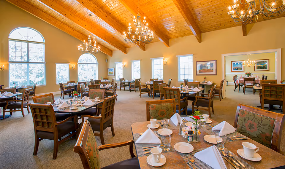 A spacious dining room with wooden vaulted ceilings and large arched windows letting in natural light. Multiple tables are set with white napkins, cups, plates, and silverware. The room features chandeliers and framed artwork on the walls, creating a warm and inviting atmosphere.