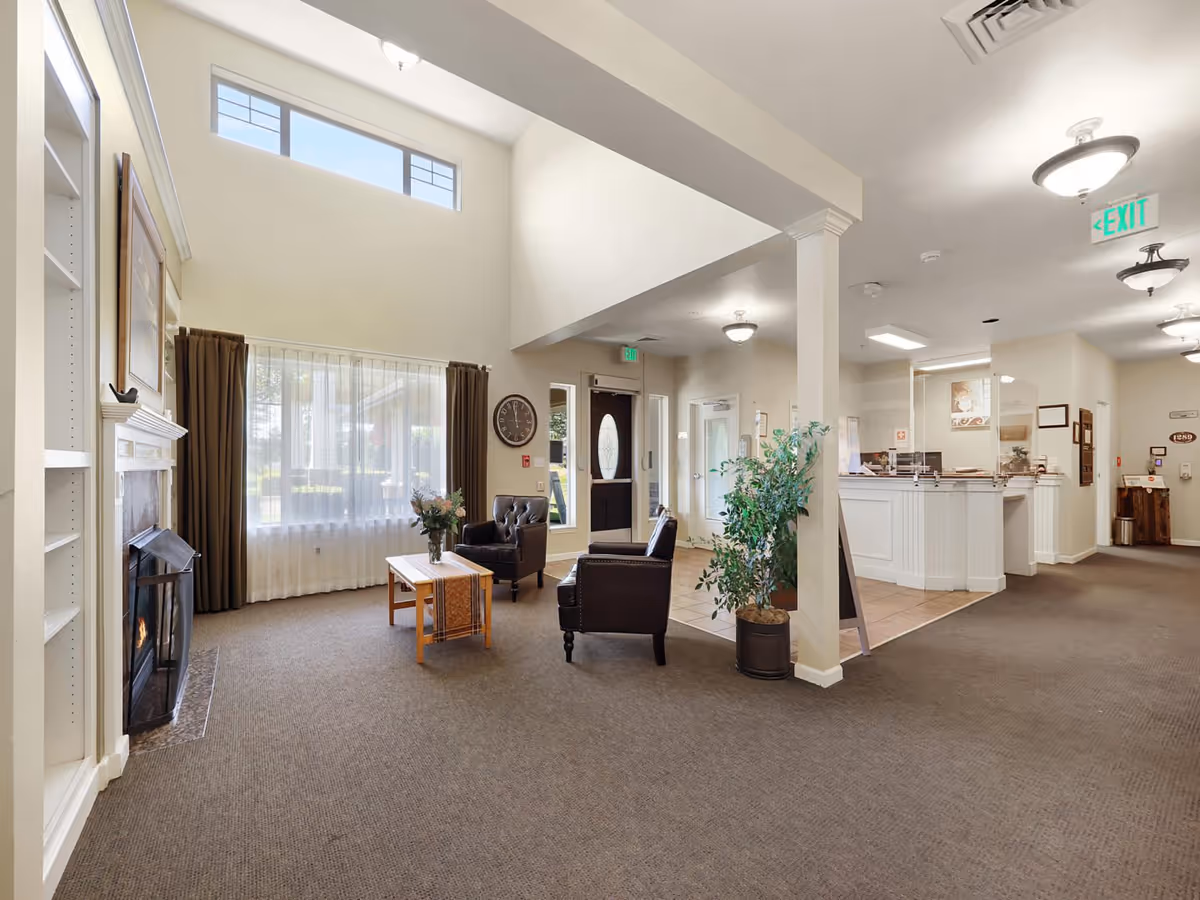 A bright and spacious interior common area with high ceilings, featuring a cozy seating arrangement with two leather armchairs and a wooden coffee table in front of a fireplace. Large windows with sheer curtains allow natural light to fill the room. To the right, there is a reception desk area with a protective screen and an exit door with a green exit sign above it. The space is decorated with a potted plant and wall clock.
