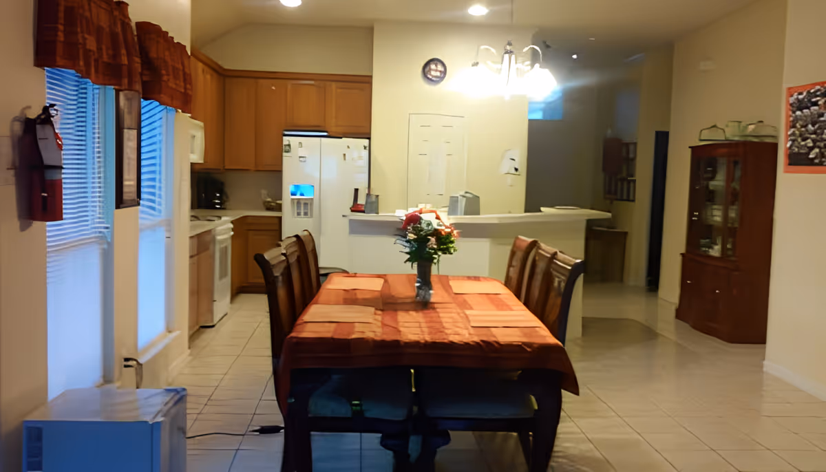 Interior view of a dining area and kitchen in a residential care facility. The dining table is covered with a red tablecloth and has a flower vase centerpiece. The kitchen features wooden cabinets, a white refrigerator, and a stove. There is a window with blinds and a valance on the left side, and a wooden cabinet with glass doors on the right side. The floor is tiled, and a ceiling light fixture is visible above the dining table.