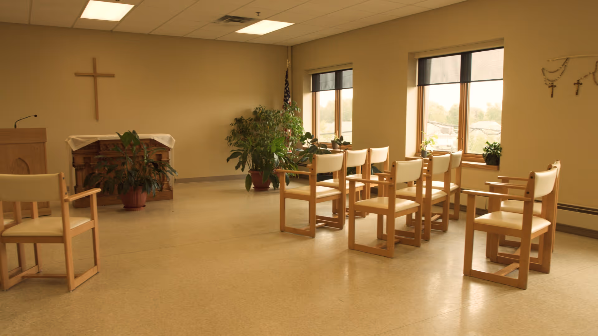 Small chapel-style gathering room with wooden chairs arranged in rows, a lectern and altar with a cross on the wall, potted plants, and windows.