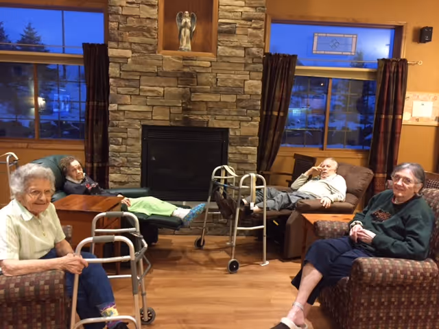 Four elderly individuals sitting and reclining in a cozy living room with a stone fireplace in the center. Two walkers are positioned near the individuals. The room has wooden floors, large windows with dark curtains, and warm lighting.