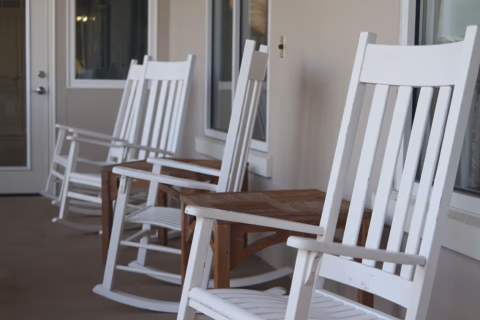 A row of white wooden rocking chairs lined up on a covered porch with a beige wall and windows in the background.