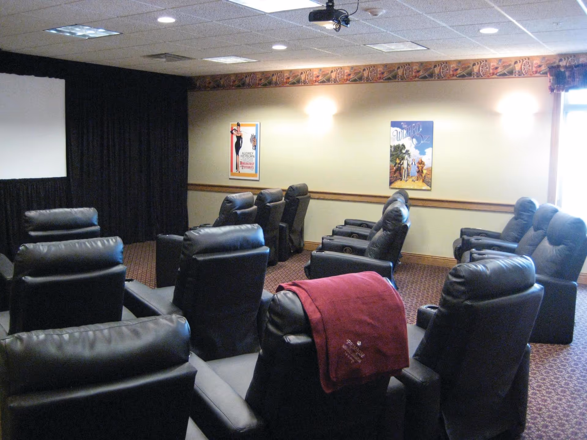 A small movie theater room with multiple rows of black leather recliner chairs facing a blank projection screen. The walls are beige with a decorative border near the ceiling and two framed movie posters. One recliner has a red blanket draped over it.