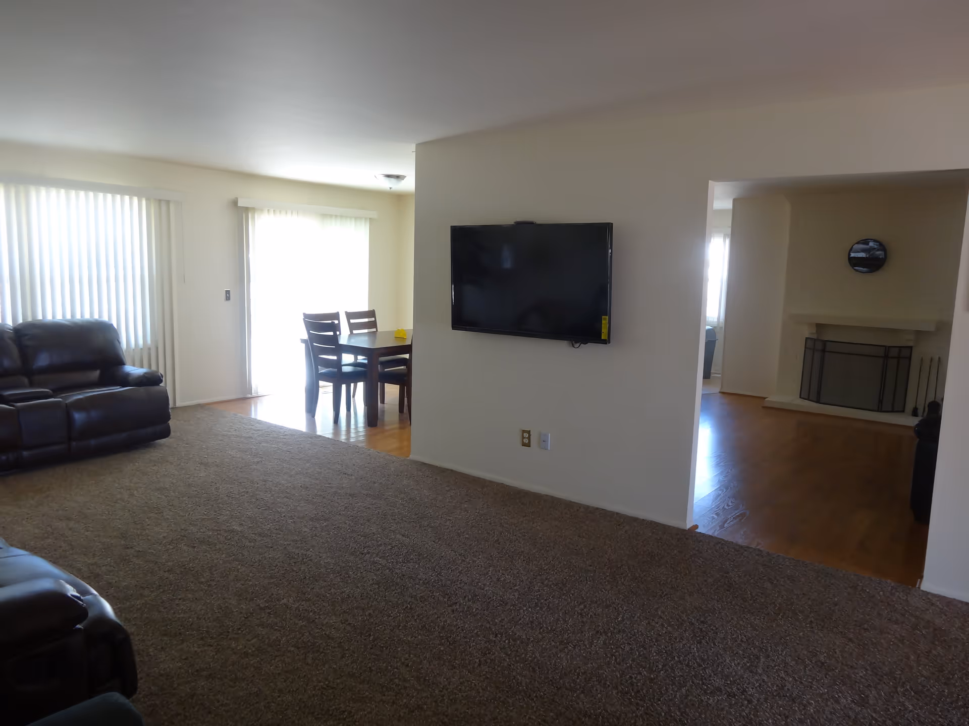 Interior view of a senior living facility showing a living room area with brown carpet and leather recliner chairs, a wall-mounted flat screen TV, and a dining area with a wooden table and chairs near large windows with vertical blinds. There is also a fireplace visible in an adjoining room with hardwood flooring.