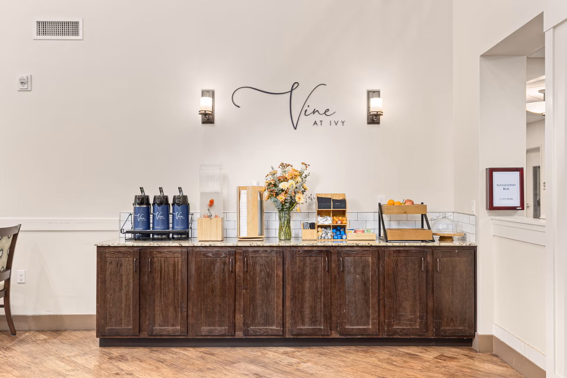 A beverage and snack station with dark wooden cabinets and a granite countertop. On the countertop are three coffee dispensers labeled 'Vine', a water dispenser with fruit inside, a container with disposable cups, a vase with flowers, compartments holding creamers and sugar packets, a fruit basket, and a covered cake stand. Above the station is a wall sign that reads 'Vine AT IVY' with two wall-mounted lights on either side. To the right, there is a suggestion box mounted on the wall.