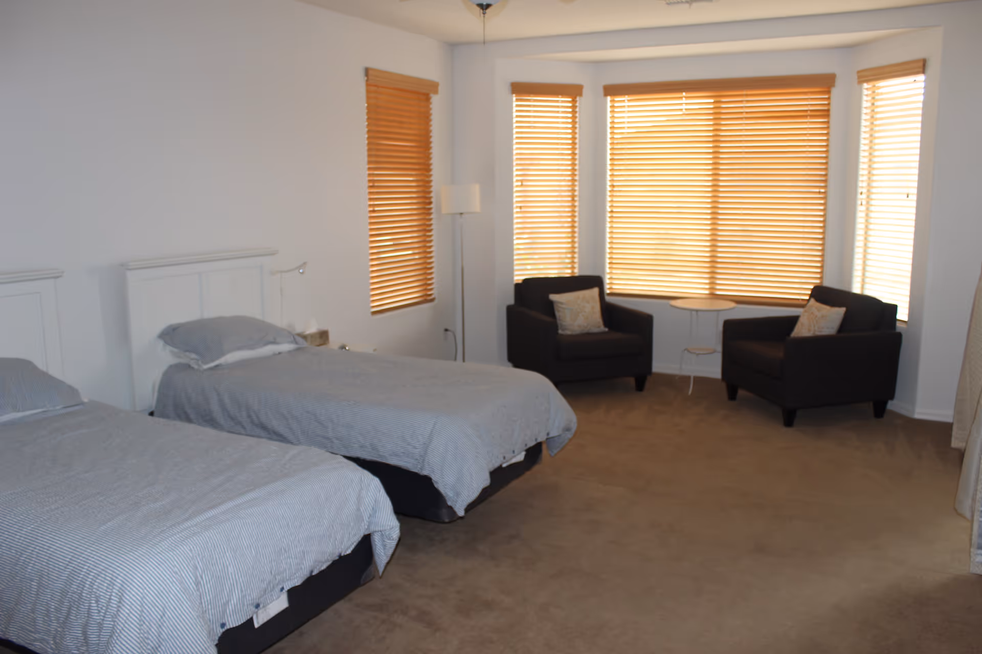 A bedroom with two single beds covered in gray striped bedding, positioned side by side. There are three windows with wooden blinds letting in natural light. In the corner near the windows, there are two dark armchairs with patterned cushions and a small round white table between them. A floor lamp stands next to one of the windows.