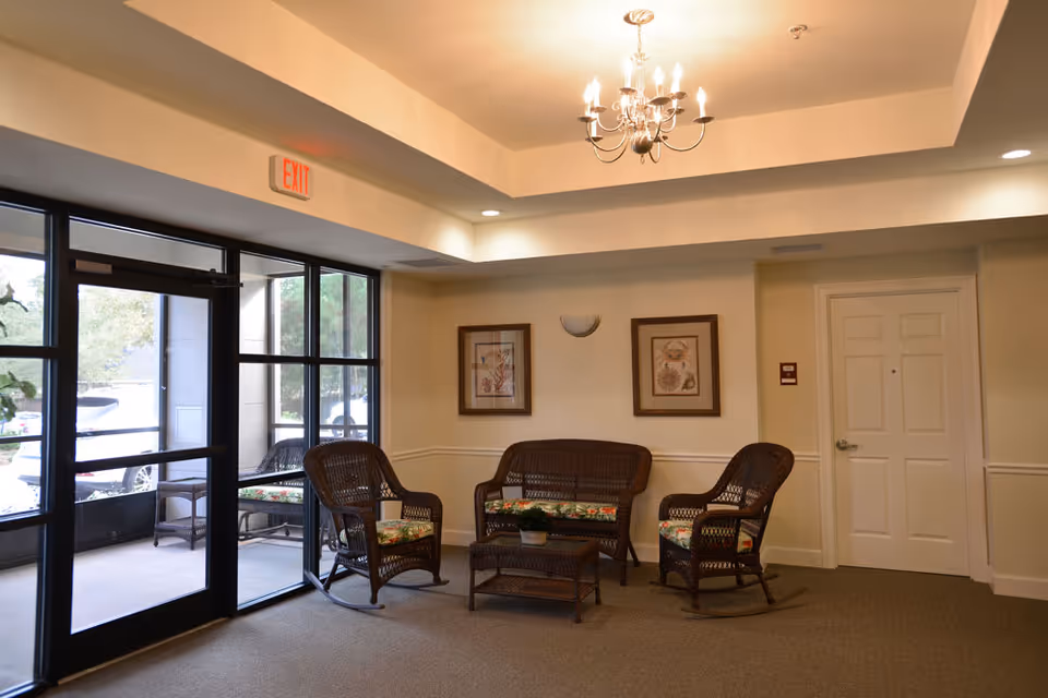 A cozy seating area inside a senior living facility with wicker furniture including two chairs, a loveseat, and a coffee table with a small plant. The room has beige walls, two framed pictures, a chandelier, and a glass door leading outside.