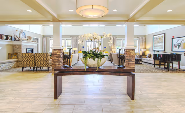 Spacious and well-lit living room area with a wooden table in the foreground holding a large decorative bowl with white orchids. The room features multiple seating areas with patterned and plain sofas, a fireplace on the left, large windows with curtains, and a black grand piano on the right. The ceiling has recessed lighting and a central hanging light fixture.
