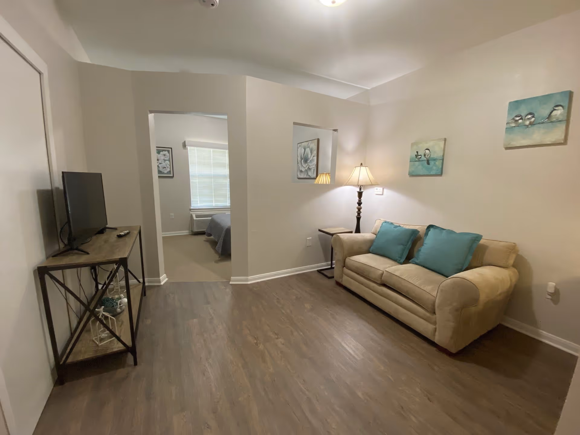 A cozy living room with a beige couch adorned with two teal pillows, a floor lamp beside it, and three bird-themed paintings on the wall. Across from the couch is a TV on a wooden stand with metal legs. The room has wooden flooring and light-colored walls. An open doorway leads to a bedroom with a bed and a window with blinds.
