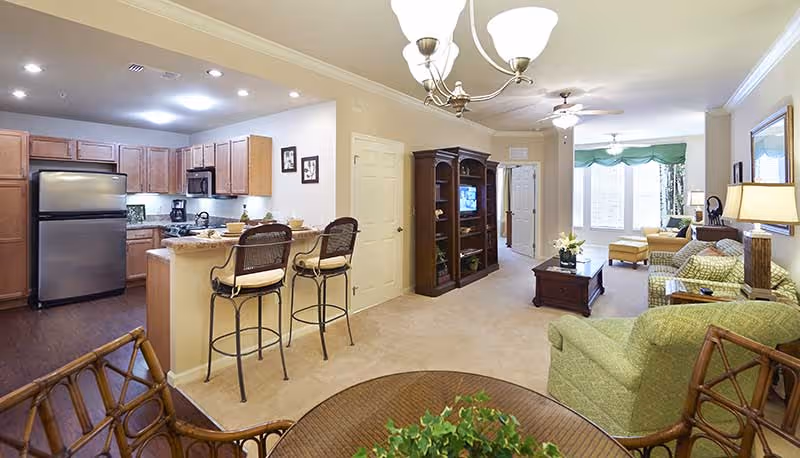 A spacious and well-lit interior of a senior living facility unit showing an open kitchen with wooden cabinets, stainless steel refrigerator, and a breakfast bar with two high chairs. Adjacent to the kitchen is a living room area with green upholstered chairs, a wooden coffee table, a TV cabinet, and large windows letting in natural light. The room has beige carpeting and light-colored walls with ceiling fans and a chandelier.