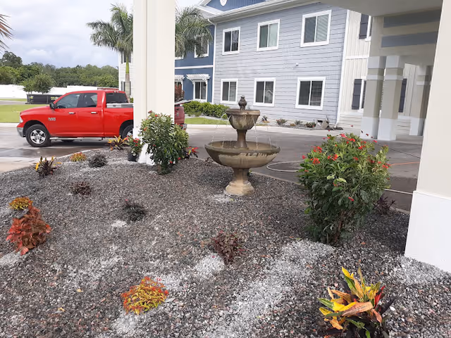 A two-tier stone fountain sits in a gravel planting bed beneath the covered entrance of a blue-gray building, with a red pickup truck and palm trees visible nearby.