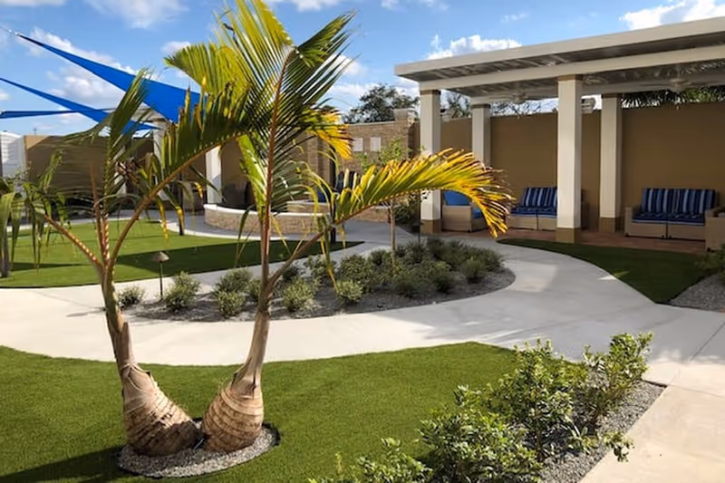 Outdoor courtyard area with a curved concrete walkway, green grass, small palm trees, and shrubs. There are shaded seating areas with blue striped cushions under a pergola structure. The sky is partly cloudy.